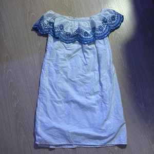 Vit klänning med blå broderad volang - Charmig vit klänning från H&M med en vacker blå broderad volangdetalj runt axlarna. Klänningen är axelbandslös och har en avslappnad passform, perfekt för varma dagar.