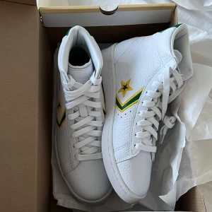 Vita sneakers med gröna och gula detaljer - Ett par klassiska Converse PRO Leather höga skor. Helt nya sneakers som hyllar basketikonen Dr. Julius Erving mannen som flög genom luften långt innan Jordan. Han bar just den här modellen på 70-talet och blev en av de mest inflytelserika afroamerikanska spelarna i NBA:s historia