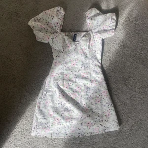 Somrig klänning  - Söt blommig klänning med puffiga kortärmar och en v-ringad design. Klänningen har blommönster i rosa, lila och grönt på en vit bakgrund. Perfekt för sommarens alla tillfällen!