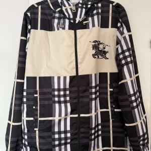 Rutig vindjacka från Burberry - Snygg rutig vindjacka från Burberry i svart och beige med dragkedja och huva. Jackan har ett stort Burberry-tryck på ryggen och en broderad detalj på framsidan. Perfekt för en stilren look.