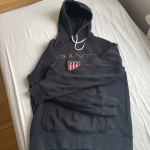 Svart hoodie från GANT - Säljer en stilren svart hoodie från GANT med ett broderat emblem på bröstet. Tröjan har en klassisk känguruficka och justerbar huva med snören. Perfekt för en avslappnad stil.