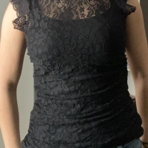 Svart spetsblus - Snygg svart spetsblus med korta ärmar och ett elegant mönster. Blusen har en hög hals och är tillverkad i ett genomskinligt spetsmaterial. Det finns linne under! Kvaliten är super bra, aldrig använt. Storlek är mellan 36/38