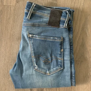 Blåa Replay Anbass jeans i storlek 29 - Snygga blå jeans från Replay med en klassisk femficksdesign och slitna detaljer på framsidan. De har en slim passform och är perfekta för en avslappnad stil. Märkeslogga på bakfickan och en läderpatch vid midjan.