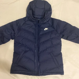 Mörkblå dunjacka från Nike - Säljer en snygg mörkblå dunjacka från Nike med huva och dragkedja. Jackan har en klassisk pufferdesign och är perfekt för kyligare dagar. Den har en broderad Nike-logga på bröstet och är både stilren och funktionell.