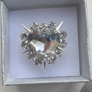 Silverfärgad ring med hjärtformad kristall - En glittrig silverfärgad ring med en stor hjärtformad kristall i mitten, omgiven av mindre kristaller och spetsiga detaljer. Perfekt för att ge en extra touch av glamour. Älskar!! ❤️Aldrig använd