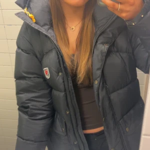 Svart dunjacka från Fjällräven - Säljer en svart dunjacka från Fjällräven med en mysig huva och dragkedja. Jackan har en klassisk design med Fjällrävens logotyp på bröstet och är perfekt för kyligare väder. Säljer den för jag vill köpa en annan jacka 