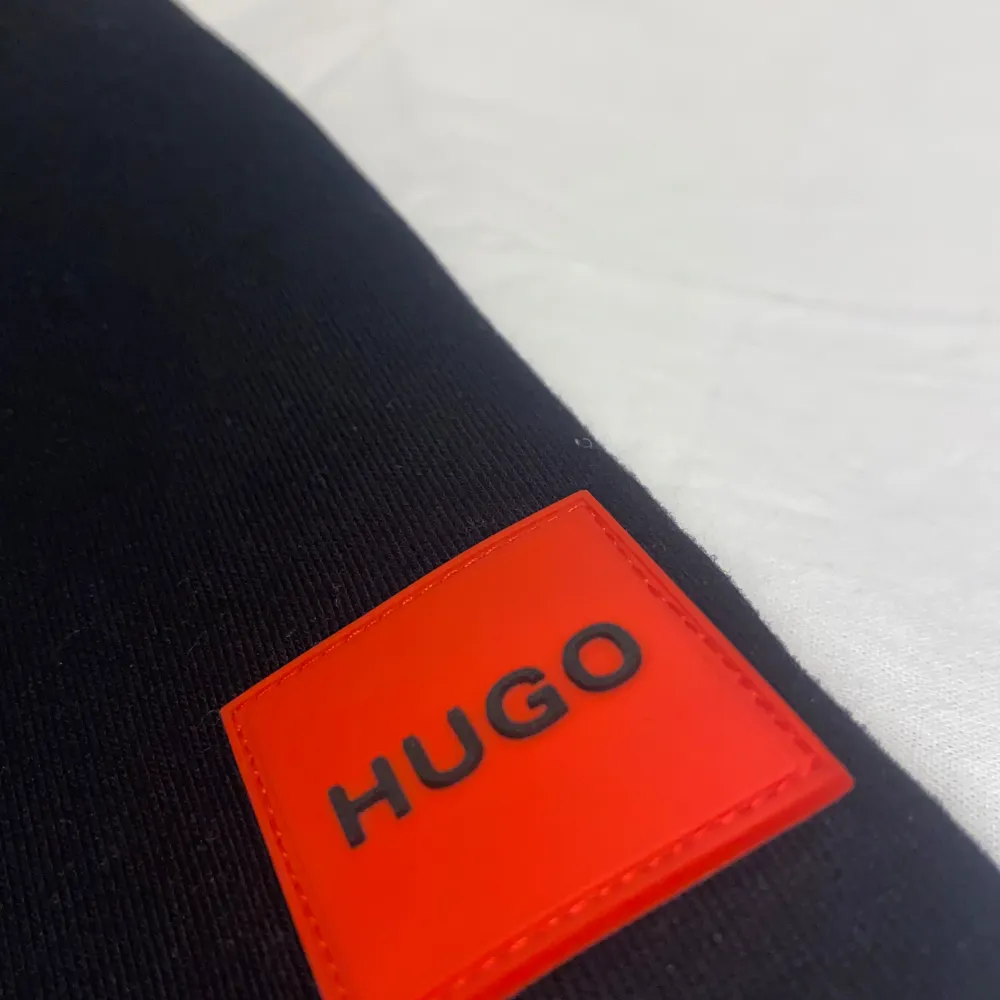 Säljer en stilren svart hoodie från Hugo med en stor logotyp i rött och vitt på bröstet. Hoodien har en justerbar huva med svarta snören och röda detaljer. Perfekt för en avslappnad stil.. Neuletakit & Villapaidat.