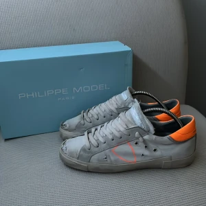 Vita sneakers från Philippe Model - Snygga grå sneakers från Philippe Model med orange detaljer. Skorna har en rund tå och snörning framtill. Tillverkade i skinn med en cool, sliten look. Perfekta för en avslappnad stil.