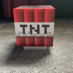 Minecraft TNT-lampa - Säljer en cool Minecraft-inspirerad TNT-lampa. Den är kubformad med ett pixelmönster i rött, svart och grått. Perfekt för spelälskare som vill ha en unik inredningsdetalj. Lampan har en vit botten med fyra svarta fötter för stabilitet.