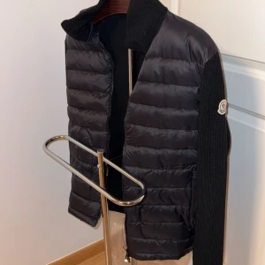 Svart quiltad jacka från Moncler - Snygg svart quiltad jacka från Moncler med stickade ärmar och krage. Jackan har en dragkedja framtill och Moncler-logotypen på ärmen. Perfekt för kyligare dagar.