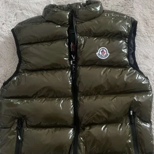 Olivgrön dunväst från Moncler - Snygg olivgrön dunväst från Moncler med glansig yta och dragkedja framtill. Västen har en hög krage och två praktiska fickor med dragkedja. Perfekt för kyliga dagar när du vill hålla stilen. Passar bra till sommaren oftast