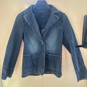 Jeansjacka med knappar - Snygg jeansjacka från H&M i klassisk blå denim. Jackan har långa ärmar och stängs med två lila knappar framtill. Den har två stora fickor fram och en stilren krage. Perfekt för en avslappnad look.