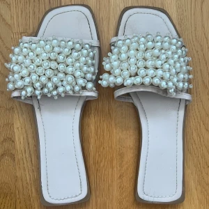 Vita sandaler med pärlor - Eleganta vita sandaler dekorerade med pärlor på ovandelen. De har en fyrkantig tå och en stilren design som passar perfekt för sommarens festligheter.