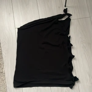 Svart oneshoulder topp med rosetter - Snygg svart oneshoulder topp med dekorativa rosetter längs sidan. Perfekt för en stilren och elegant look. Tillverkad i ett mjukt och bekvämt material.