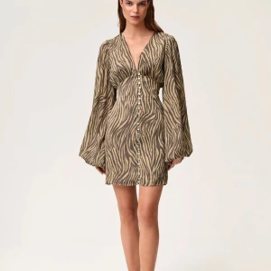 Adoore klänning zebra - Trendig zebramönstrad klänning med v-ringning och knappar framtill. Klänningen har långa ärmar och en kort längd, perfekt för en stilfull look. Helt ny