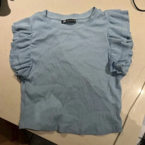 Ljusblå ribbad topp från Zara - Säljer en söt ljusblå ribbad topp från Zara med volangdetaljer på ärmarna. Toppen har en rund halsringning och korta ärmar. 