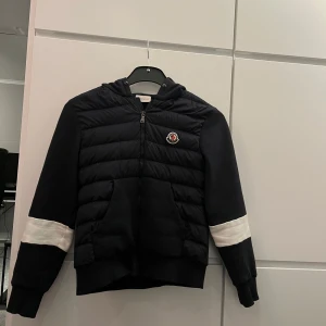 Svart quiltad jacka från Moncler - Snygg svart quiltad jacka från Moncler med huva och dragkedja. Jackan har vita detaljer på ärmarna och en broderad logotyp på bröstet.
