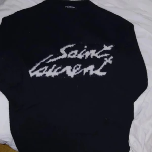 Svart tröja från Saint Laurent - Säljer en svart tröja från Saint Laurent med vit text på framsidan. Tröjan har en klassisk design och är perfekt för en stilren look. Passar bra till både jeans och kjol. Använd endast en gång och har I get tecken  på användning