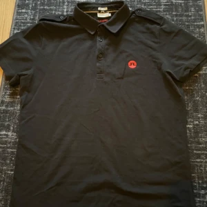 Svart pikétröja från J.Lindeberg - Säljer en stilren svart pikétröja från J.Lindeberg med röd logga på bröstet. Tröjan har korta ärmar och en klassisk krage med knappar. Perfekt för en avslappnad men snygg look.