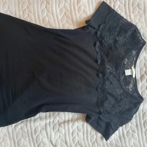 Svart spetsdetaljerad topp från H&M - Snygg svart topp från H&M med spetsdetaljer över axlarna och bröstet. Toppen har korta ärmar och en rund halsringning. Perfekt för en stilren look.