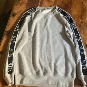 Grå sweatshirt från Valentino - Säljer en stilren grå sweatshirt från Valentino med svarta band längs ärmarna där 'VLTN' är tryckt i vitt. Tröjan har en klassisk passform med rund hals och långa ärmar. Perfekt för en avslappnad och trendig look. Väldigt bra skick! Inga fläckar eller slitage. Kom pm om du har fler frågor