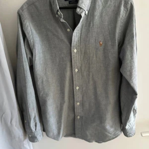 Grå skjorta från Ralph Lauren - Säljer en stilren grå skjorta från Ralph Lauren med klassisk krage och broderad logga på bröstet. Skjortan har långa ärmar och knappar framtill. Perfekt för en snygg och avslappnad look.                             Priset är kan diskuteras vid snabb affär 