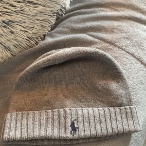 Grå stickad mössa från Ralph Lauren - Snygg beige stickad mössa från Ralph Lauren med broderad logga framtill. Perfekt för kyliga dagar och ger en stilren look. Passar både till vardags och mer uppklädda tillfällen.