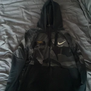 Svart och grå camo hoodie från Nike - Säljer en snygg svart och grå camo hoodie från Nike med Dri-FIT-teknologi. Hoodien har en dragkedja framtill och en stor huva. Perfekt för en sportig look eller avslappnade dagar.