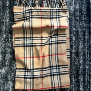 Beige rutig halsduk från Burberry  - Snygg beige halsduk med klassiskt rutmönster i svart, vitt och rött. Perfekt för att hålla sig varm och stilren under kyliga dagar. Halsduken har fransar i ändarna som ger en extra touch av elegans.