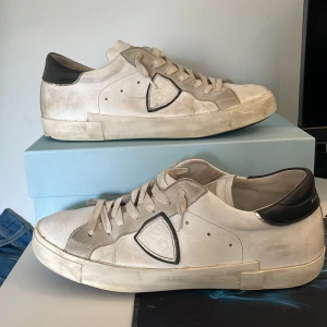 Vita phillipemodels storlek 43 skick 7/10  - Snygga vita sneakers med svarta detaljer och snörning. Skorna har en rund tå och en stilren design som passar perfekt till vardags. De har en kontrasterande svart häl och en unik logotyp på sidan.