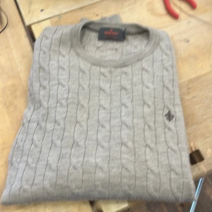 Grå  tröja från Morris - Säljer en stilren grå kabelstickad tröja från Morris i 100% merinoull. Tröjan har en diskret logga på bröstet och rund halsringning. Perfekt för kyliga dagar!