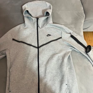 Grå Nike tech Fleece - Säljer en stilren grå huvtröja från Nike med dragkedja och svart logo på bröstet. Perfekt för en sportig look!