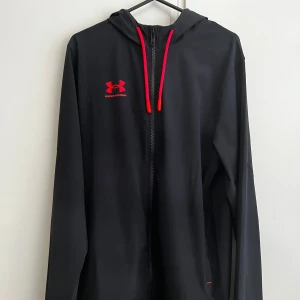 Svart vindjacka från Under Armour - Snygg svart vindjacka från Under Armour med röd logga och dragkedja. Jackan har en huva och långa ärmar, perfekt för träning eller vardagsbruk. Den är tillverkad i ett lätt och bekvämt material.