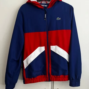 Blå och röd vindjacka från Lacoste - Säljer en snygg blå och röd vindjacka från Lacoste Sport. Jackan har en dragkedja framtill och en huva med röd insida. Den har elastiska muddar och en vit detalj på framsidan. Perfekt för sportiga aktiviteter eller en casual look.