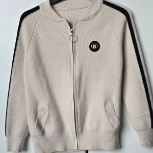 Beige stickad kofta med dragkedja - Säljer en beigevit stickad kofta med dragkedja och svarta ränder längs ärmarna. Storlek och märke står inte, men skulle anta att det är storlek S/36. (Kontrollera mått) Tröjans högra arm har även en svart liten fläck men påverkar inte kvaliten. 