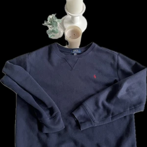 Mörkblå tröja från Ralph Lauren - Säljer en stilren mörkblå tröja från Ralph Lauren med det klassiska röda logotypbroderiet på bröstet. Tröjan har långa ärmar och en rund halsringning. Perfekt för en avslappnad och snygg look. Lite nopprig annars väldigt bra!