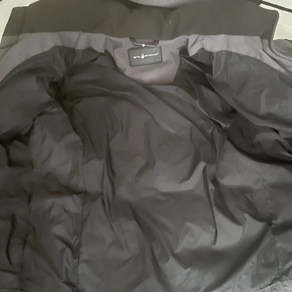 Snygg och varm grå dunjacka från Sail Racing med huva och dragkedja. Perfekt för kyliga dagar. Jackan har en stilren design med flera praktiska fickor.. Takit.