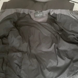 Grå dunjacka från Sail Racing - Snygg och varm grå dunjacka från Sail Racing med huva och dragkedja. Perfekt för kyliga dagar. Jackan har en stilren design med flera praktiska fickor.