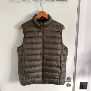 Dunväst Uniqlo - Säljer en olivgrön dunväst från Uniqlo. Västen är lätt och har en dragkedja framtill. Perfekt för lager-på-lager under kyliga dagar. Den har en stilren design med hög krage. Aldrig använd