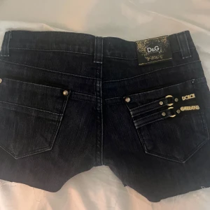 Vintage jeansshorts från Dolce & Gabbana - Snygga svarta jeansshorts från Dolce & Gabbana med guldfärgade detaljer och broderad logga. Shortsen har en låg midja och fransiga kanter för en trendig look. Perfekta för sommarens alla äventyr!