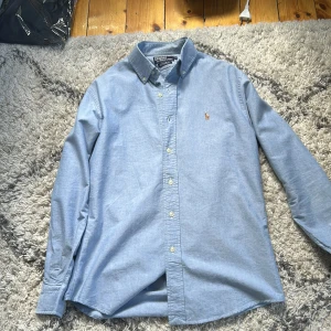 Blå skjorta från Polo Ralph Lauren - Snygg ljusblå skjorta från Polo Ralph Lauren i 100% bomull. Klassisk button-down krage, broderad logga på bröstet och långärmad modell. Custom fit som sitter riktigt bra och passar till många olika stilar. Perfekt för dig som gillar stilrena plagg med premiumkänsla.