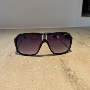 Svarta Carrera solglasögon retro - Snygga svarta Carrera solglasögon med stor, fyrkantig retroform och lila tonade glas. Tuff metall-detalj i mitten och Carrera-logga på skalmen. Ramen är i plast och glasen har en cool gradient-effekt. Perfekt för dig som gillar statement-accessoarer.