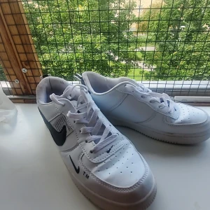 Nike Air Force 1 vita sneakers - Fräscha Nike Air Force 1 sneakers i vitt skinn med svarta detaljer och klassisk swoosh-logga på sidorna. Skorna har snörning, perforerad tå och låg profil. Perfekta för dig som gillar en clean och tidlös streetstil.