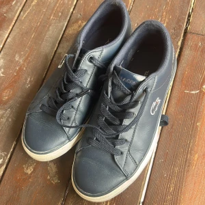 Mörkblå sneakers från Lacoste - Snygga mörkblå sneakers från Lacoste i skinn med klassisk krokodillogga på sidan och vit sula. Skorna har rund tå, snörning och diskreta sömdetaljer. Perfekta för en clean och sportig look.