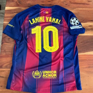 Barcelona 25/26 - Lamine Yamal #10 - Snygg FC Barcelona matchtröja med Lamine Yamal och nummer 10 på ryggen. Tröjan är kortärmad, har klassiska blå och röda ränder, gula detaljer och flera tryck som Spotify, UNHCR och Champions League. Tillverkad i lätt Dri-FIT material som andas.