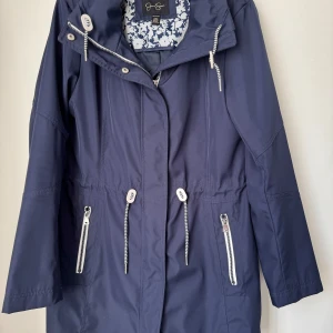 Mörkblå parkajacka med snörning - Parkajacka i mörkblått med långa ärmar, dragsko i midjan och huva. Jackan har vita detaljer på dragkedjor och snörning samt blommigt foder. Perfekt för dig som vill ha en stilren och funktionell jacka.