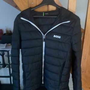Svart pufferjacka från BOSS,   - Snygg svart pufferjacka från BOSS med huva och vit dragkedja. Jackan har quiltad design, raka linjer och logga på bröstet. Perfekt för kyliga dagar och enkel att matcha med olika outfits. Lätt och smidig att bära, passar dig som gillar stilren look. Skicka mycket bra/ knappt använd. Original pris: 3123 kr.