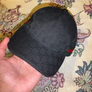 Svart Gucci keps med GG-mönster - Snygg svart keps från Gucci med diskret GG-monogram över hela tyget. Klassisk böjd skärm och justerbart spänne bak i svart skinn. Sidan har den ikoniska gröna och röda randen. Perfekt accessoar för att lyfta din outfit. Råka ta av taggen