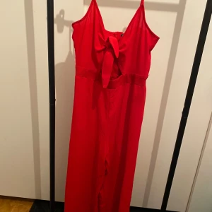 Röd jumpsuit med rosett från H&M - Snygg röd jumpsuit från H&M Divided i storlek 40. Klänningen har smala axelband, en stor rosett framtill och en fin omlott-detalj vid bysten. Materialet är mjukt och lätt, perfekt för varma dagar. Dragkedja i ryggen för enkel påklädning.