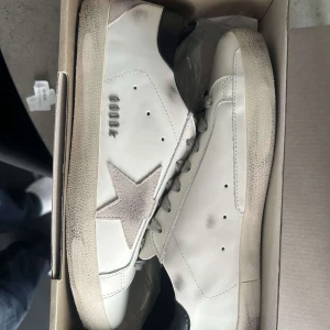 Golden Goose vita sneakers med stjärna - Snygga och trendiga Golden Goose sneakers i vitt skinn med grå mockadetaljer och ikonisk stjärna på sidan. Grå snörning, svart hälparti och GGDB-logga. Snyggt slitna detaljer och platt sula ger en avslappnad vibe. Hör av er vid fundering.